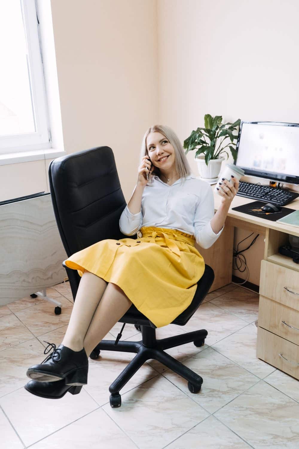 Blonde Frau in gelbem Rock, schwarze Schuhe, weiße Bluse, Kaffee und Handy in der Hand telefoniert lächelnd am Schreibtisch mit Beinen übereinandergeschlagen. Helles Büro mit Zimmerpflanze im Hintergrund.