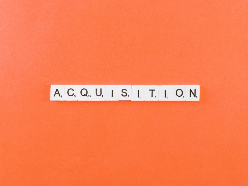 Scrabble-Würfel, die das Wort “Akquisition” bilden, auf orangem Hintergrund.
