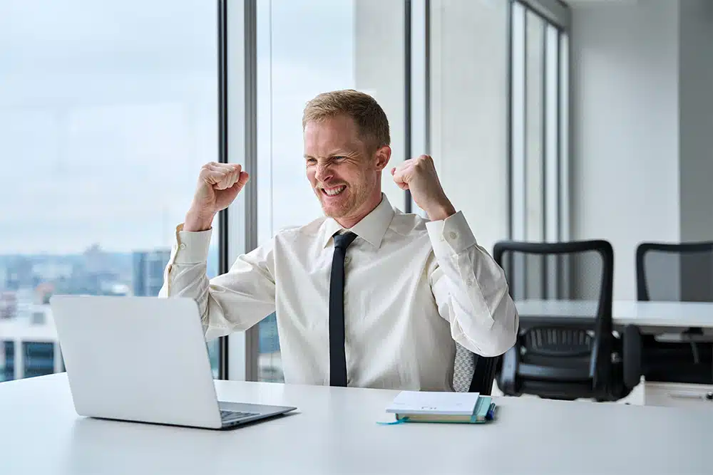 Mann im Business-Outfit jubelt vor Laptop in modernem Büro mit Fensterfront.