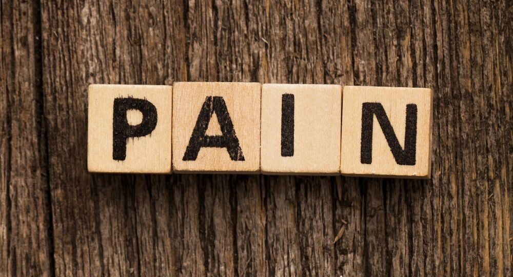 Hellbraune Würfel mit Buchstaben, eine Reihe bildet das Wort Pain, die andere darunter das Wort Gain. Liegen auf dunklem Holz.