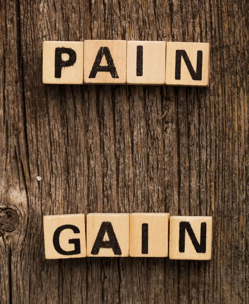 Hellbraune Würfel mit Buchstaben, eine Reihe bildet das Wort Pain, die andere darunter das Wort Gain. Liegen auf dunklem Holz.