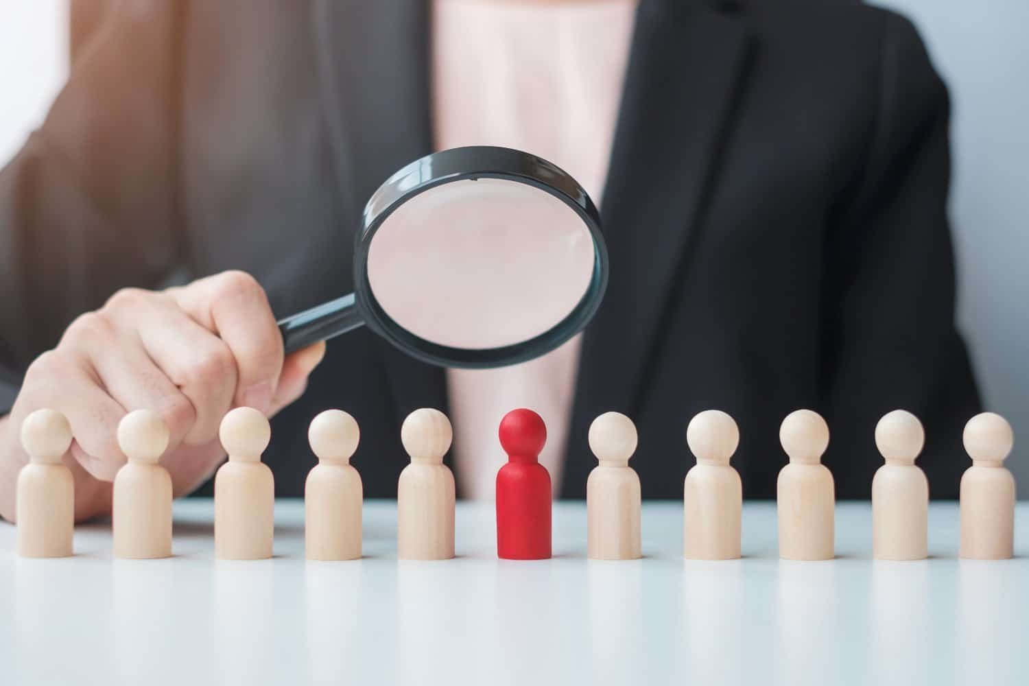 Brustbereich einer Frau in schwarzem Blazer und weißer Bluse, die vor weißem Schreibtisch sitzt und Lupe über eine rote Holzfigur in der Mitte von hellbraunen Holzfiguren hält