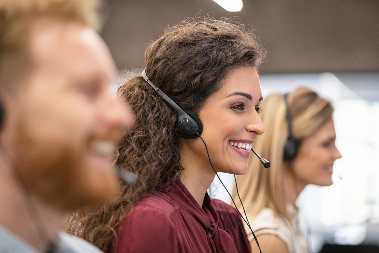 Ein Mann ganz vorne und zwei Frauen dahinter ein einer Reihe sitzen mit Headsets auf den Köpfen am Computer und telefonieren lächelnd.