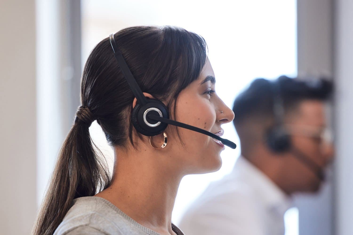 Frau und Mann im Profil sitzen mit Headset auf dem Kopf in einer Reihe vor einem Schreibtisch. Weiße Wand und Fenster leicht verschwommen im Hintergrund.