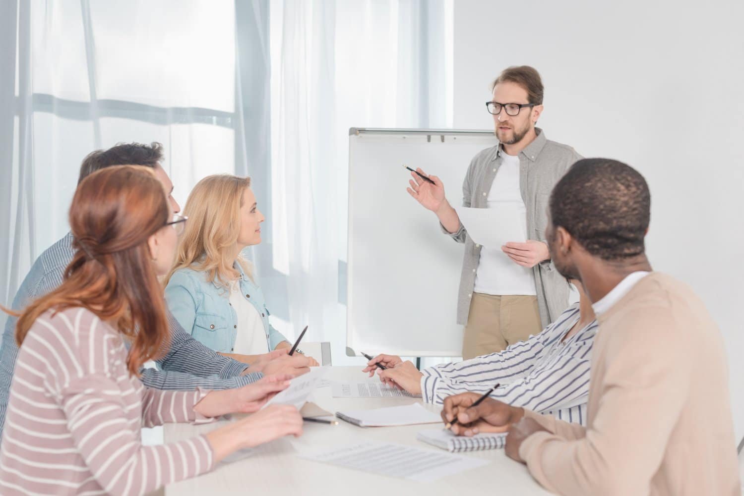 Mann mit Bart und Brille hält Papier und Stift in der Hand und steht sprechend vor Whiteboard, während fünf Personen an einem weißen Tisch mit Papier und Stift sitzen und in seine Richtung blicken. Weiße Wand, große Fenster mit dunklen Rahmen und weiße, zugezogene Gardinen im Hintergrund.