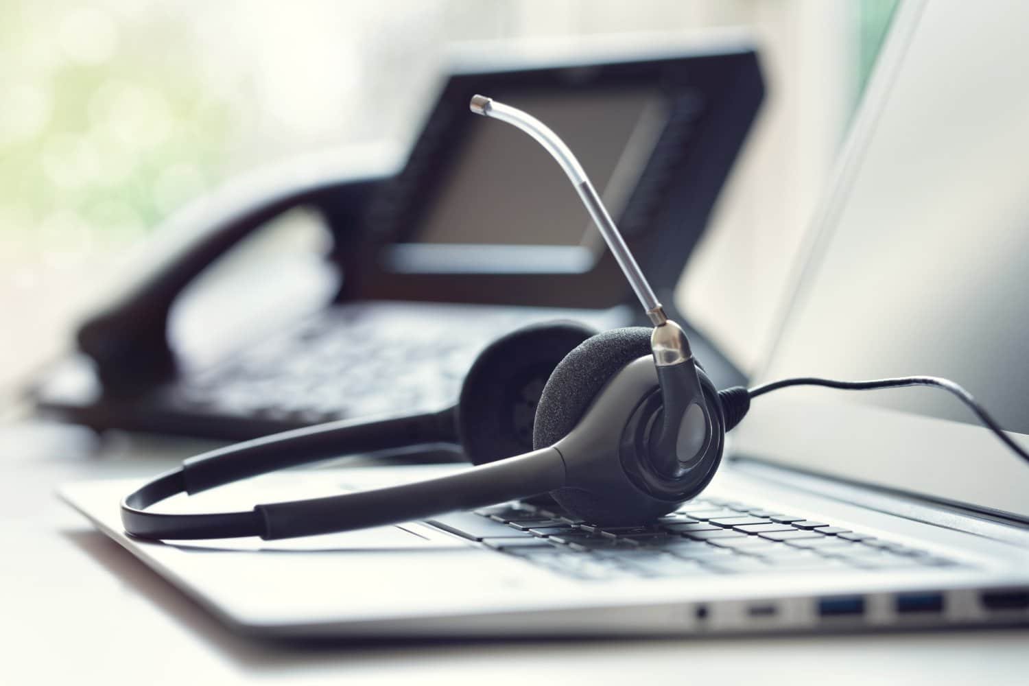 Schreibtisch mit Headset auf Laptoptastatur, links daneben Telefonanlage mit Bildschirm. Verschwommener, weißer Hintergrund.