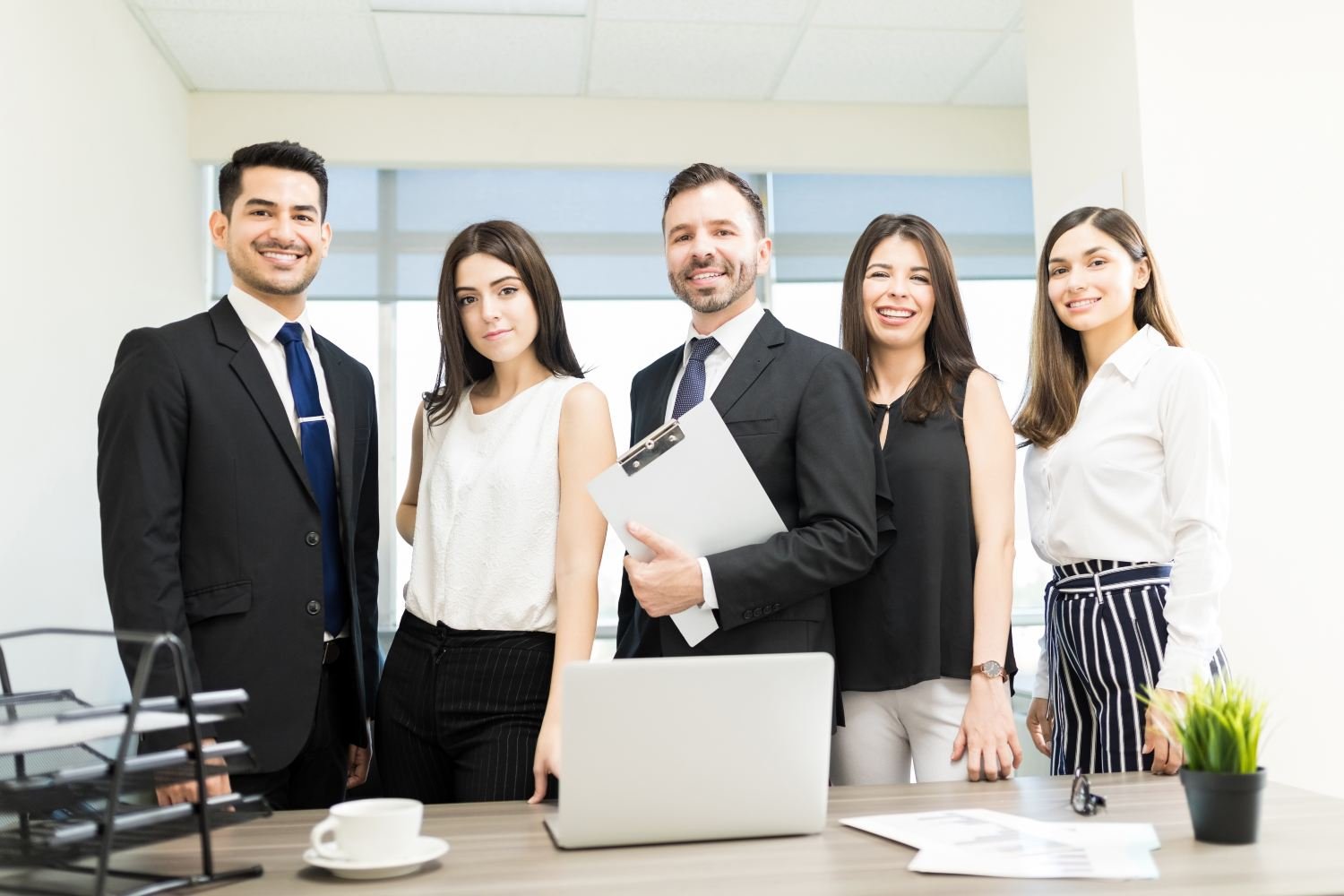 Drei Frauen und zwei Männer in einer Reihe vor Schreibtisch in schwarz-weißer Businesskleidung schauen in die Kamera, der Mann in der Mitte hält ein Klemmbrett. Laptop, Kaffeetasse, Dokumente und Zimmerpflanze auf Schreibtisch. Hohe und breite Fenster mit weißen Rahmen im Hintergrund.
